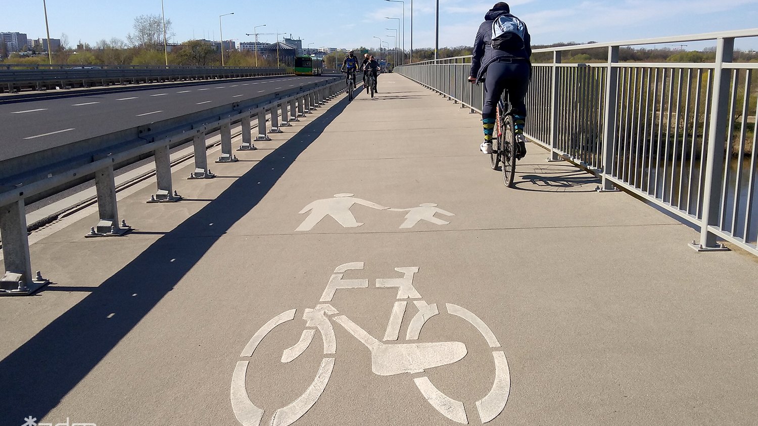 Zdjęcie drogi dla rowerzystów i pieszych, odzielonej metalową barierką, po drodze jadą rowerzyści