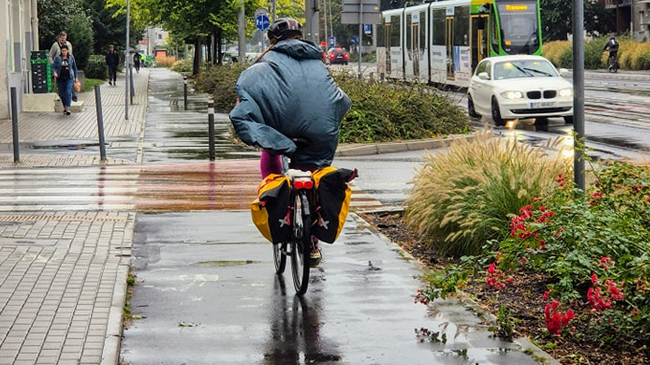 Zdjęcie, deszcz, rowerzysta jedzie w pelerynie przeciwdeszczowej po drodze rowerowej.