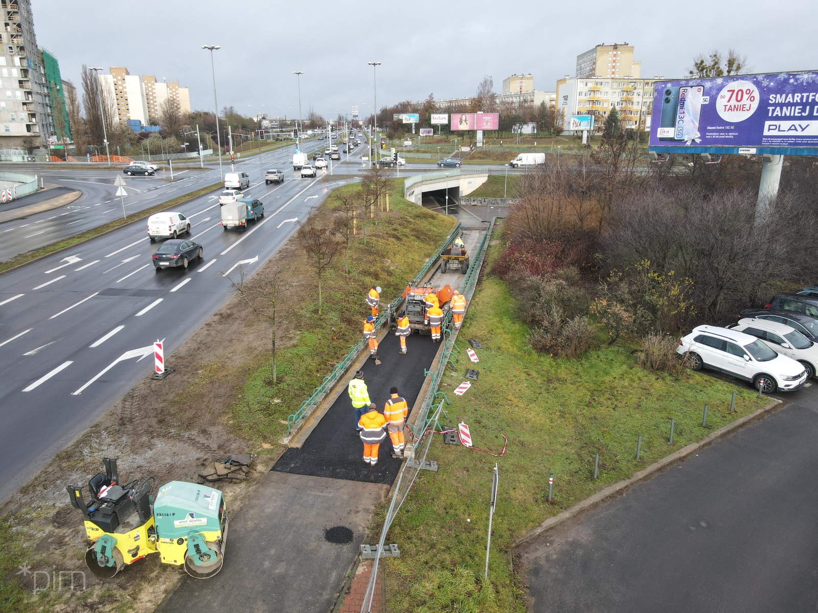 Roboty drogowe zwiazane z trasą rowerową przy przystanku PST Słowniańska. Pracownicy drogowi ubrani w stroje odblaskowe, maszyna do układania nawierzchni, obok skrzyżowanie drogowe. - grafika artykułu