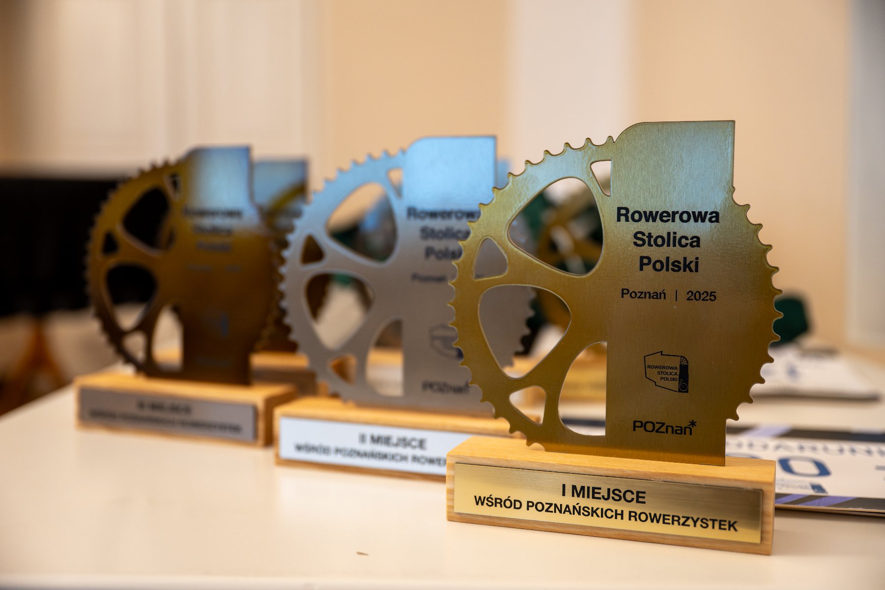 Rowerowe nagrody dla najlepszych rowerzystek i rowerzystów. Trzy statuetki w kształcie rowerowych zębatek ustawione na białym stole. Na pierwszym planie złota nagroda z napisem: "Rowerowa Stolica Polski, Poznań 2025, I miejsce wśród poznańskich rowerzystek". W tle widoczne są kolejne trofea w srebrnym kolorze.