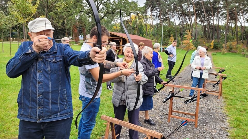 Teren zielony, w oddali drzewa. Mężczyzna strzela z kuszy. Obok młodszy mężczyzna pokazuje kobiecie, jak strzelać z kuszy. Kilka osób przygląda się temu.