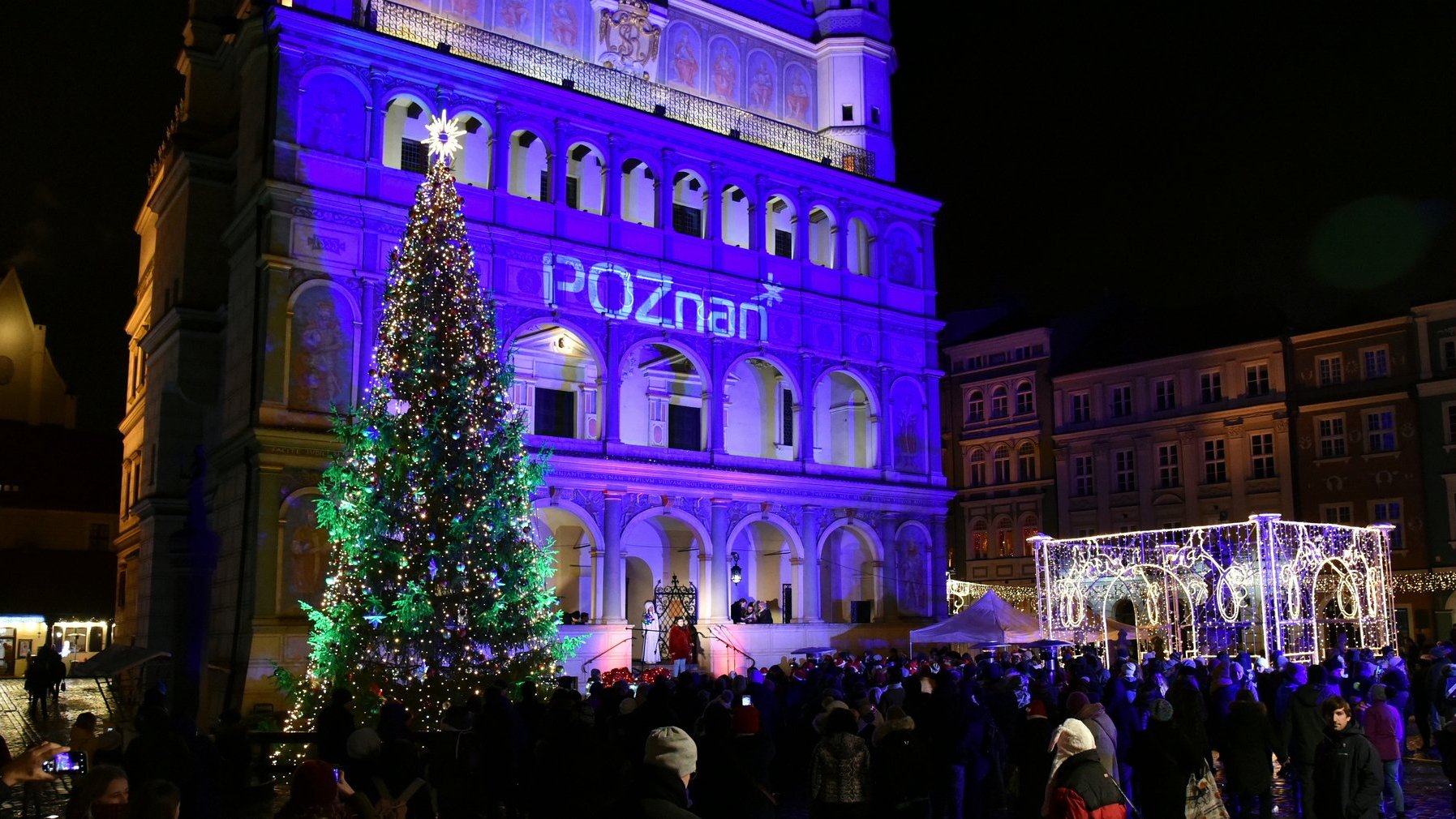 Zdjęcie przedstawia oświetlony na niebiesko Ratusz. Obok niego stoi choinka. Na zdjęciu widać też tłum ludzi oraz świąteczne iluminacje.