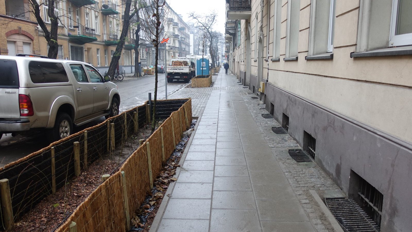 Zdjęcie przedstawia chodnik, posadzone obok rośliny oraz stojące na poboczu jezdni samochody.