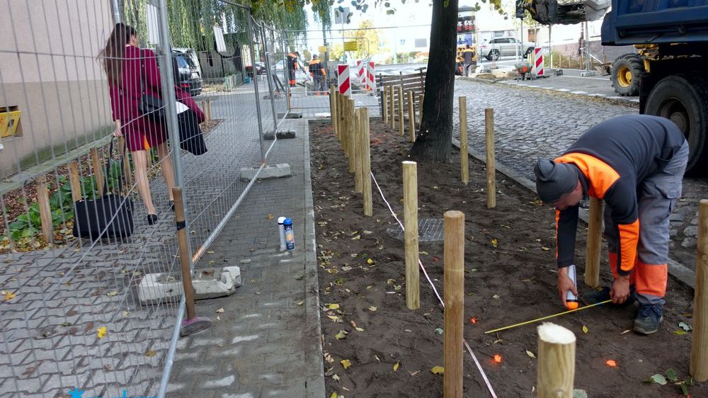 Zdjęcie przedstawia mężczyznę mierzącego odcinek pobocza oraz kobietę z walizką na kółkach idącą chodnikiem.