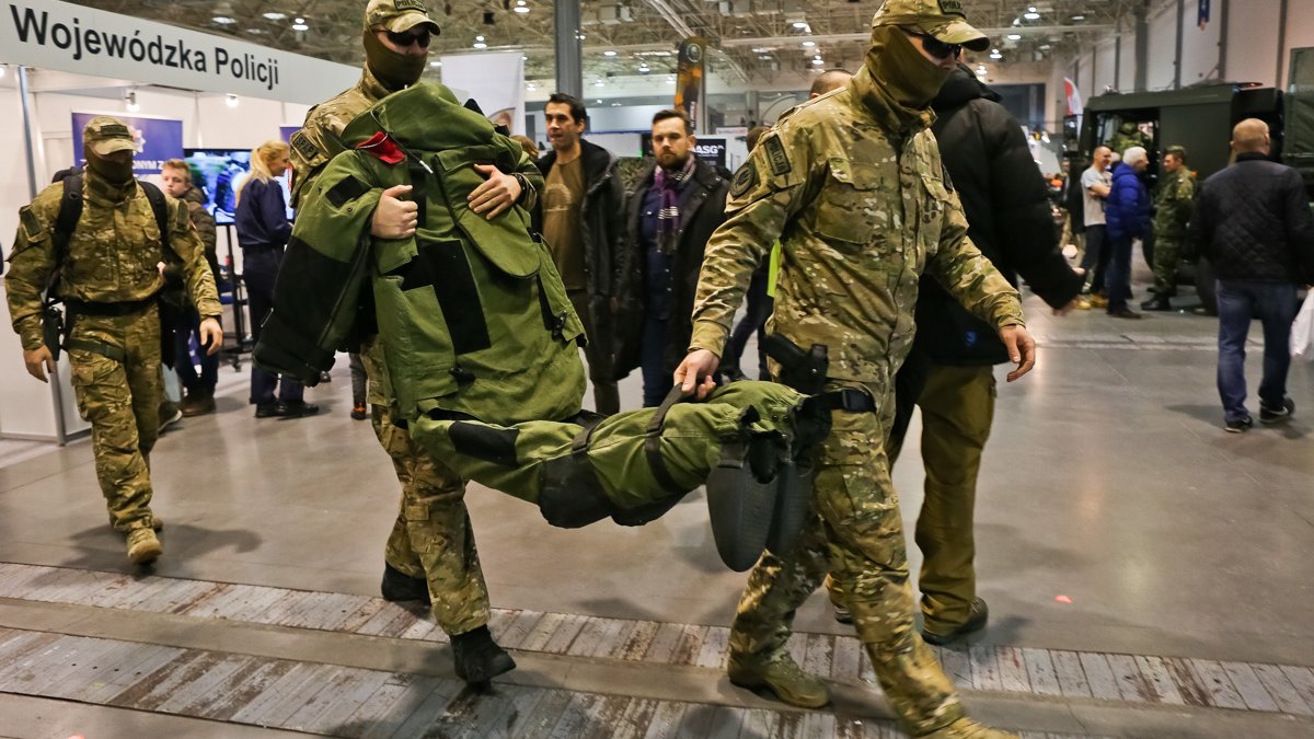 Zdjęcie przedstawia ludzi w wojskowych strojach niosących zielony kombinezon. W tle widać innych ludzi.