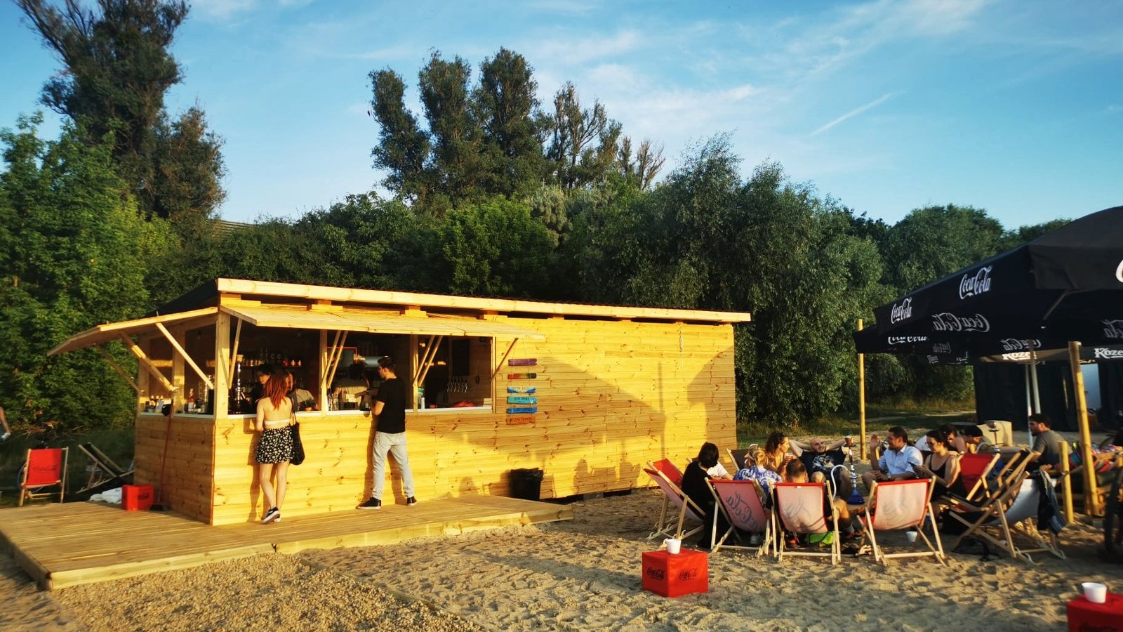 Zdjęcie przedstawia ludzi siedzących na leżakach na plaży oraz drewniany domek, w którym znajduje się bar.