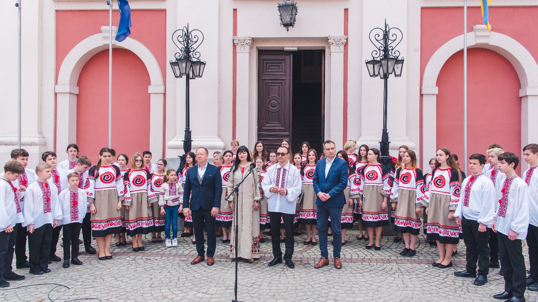 Na zdjęciu chór przed budynkiem urzędu miasta, przed artystami czworo dorosłych, przed którymi stoi mikrofon