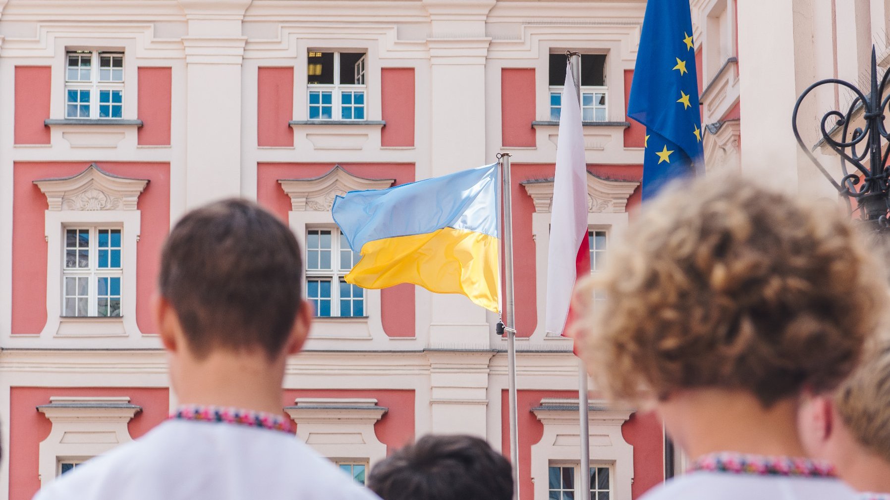 Na zdjęciu młodzi chórzyści, stojący tyłem do obiektywu, przed nimi widać trzy flagi: Ukrainy, Polski oraz Unii Europejskiej