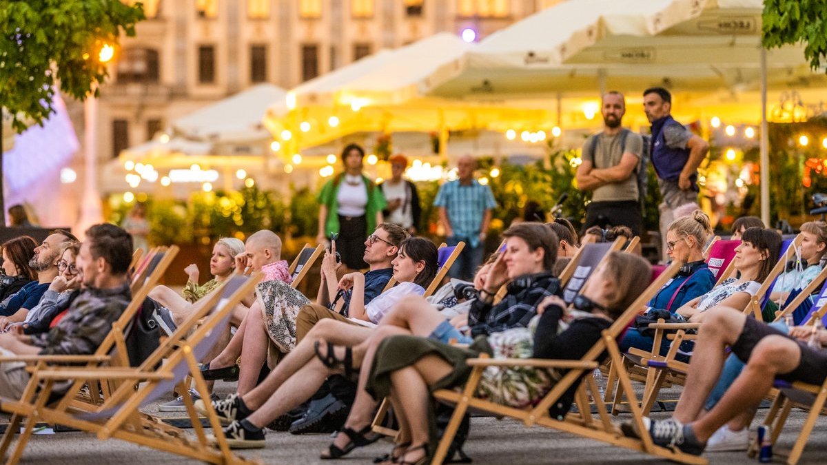 Na zdjęciu grupa ludzi leży na leżakach w centrum miasta, prawdopodobnie oglądają fim