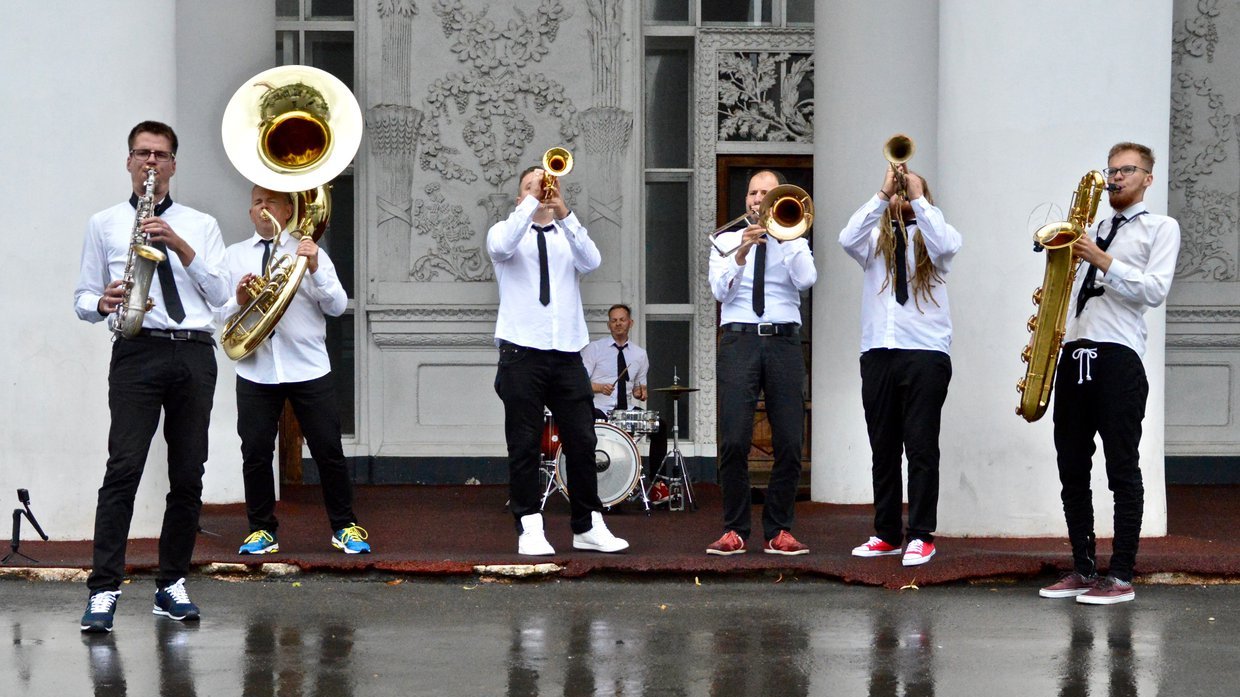 Na zdjeciu grupa 6 mężczyzn z instrumentami dętymi, w tle siedzi perkusista