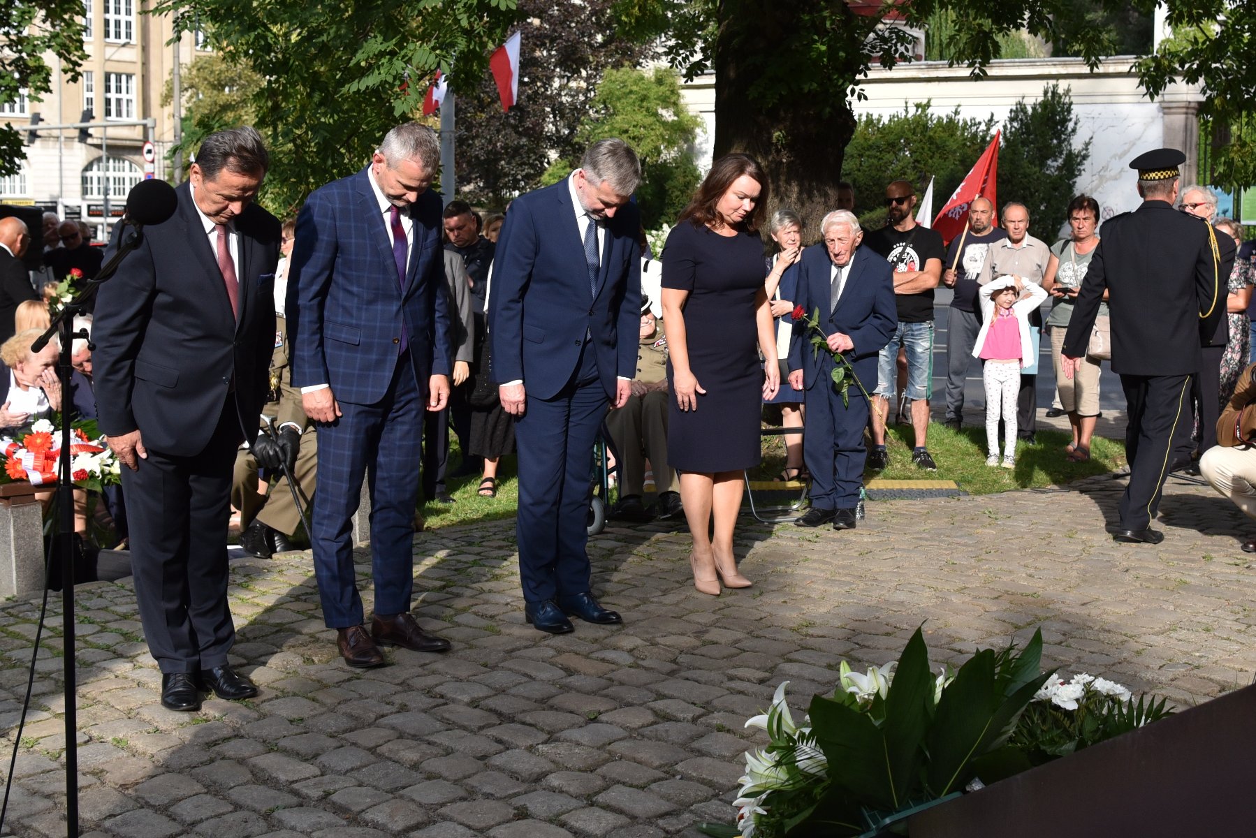 Na zdjęciu przedstawiciele władz rządu i samorządu składają wiązankę kwiatów, pochyleni - grafika artykułu