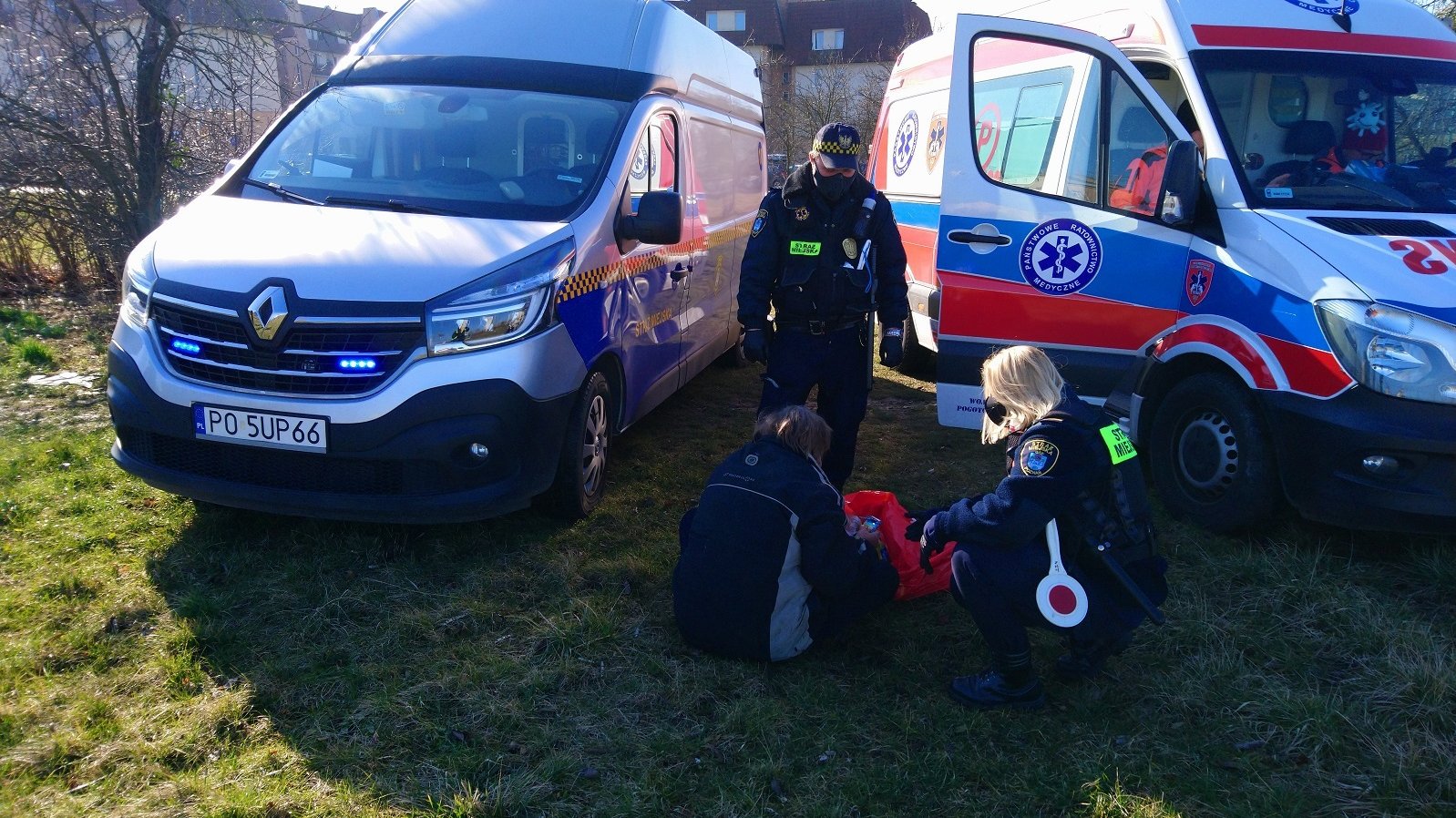 Na pierwszym planie strażnicy miejscy pomagają siedzącej na ziemi osobie. W tle radiowóz straży miejskiej i karetka pogotowia. - grafika artykułu