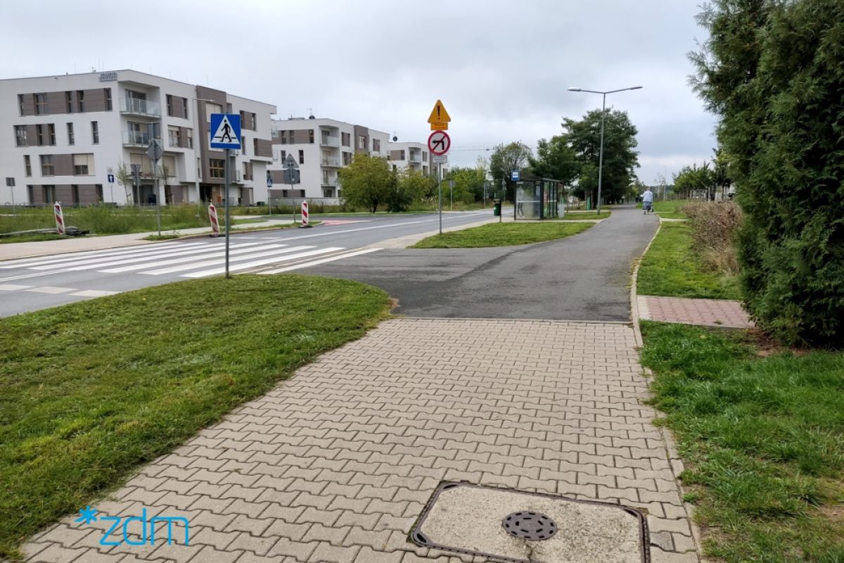 Na pierwszym planie droga rowerowa, w tle wiata przystankowa i zabudowania. - grafika artykułu