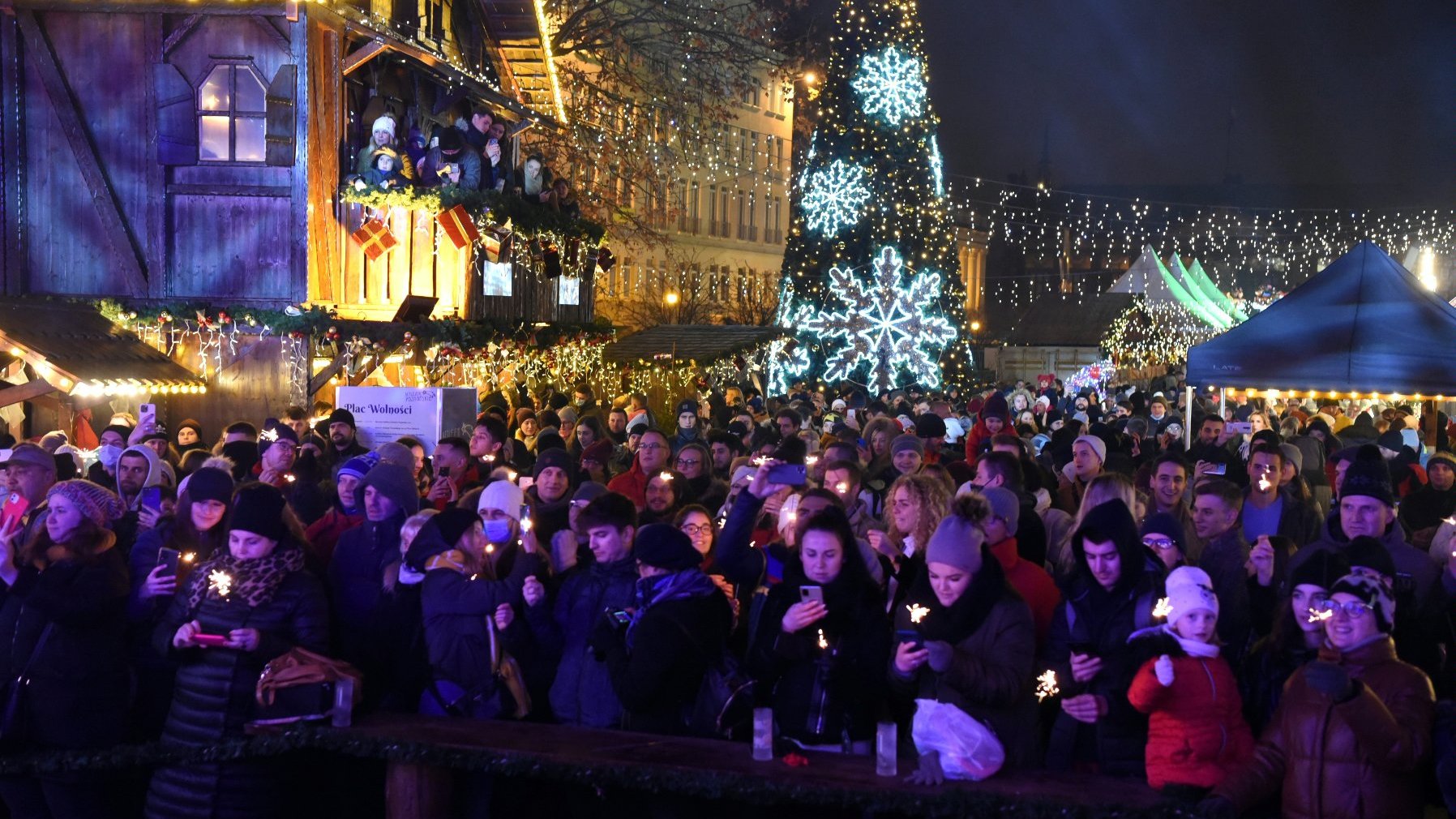 Na zdjęciu ludzie zgromadzeni na placu wieczorem, w tle choinka, iluminacje oraz stoiska jarmarku