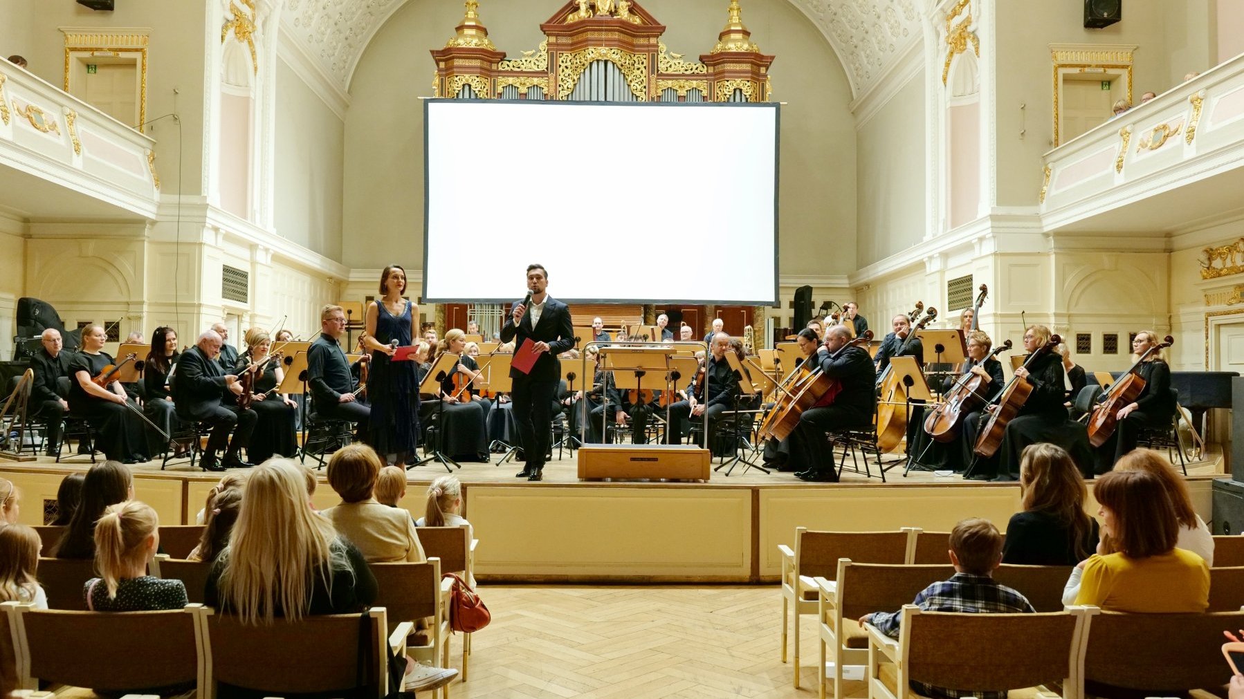 Na zdjęciu muzycy na scenie, przed nimi prowadzący, widać także publiczność