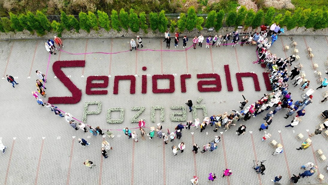 Na zdjęciu widoczny z lotu ptaka napis "Senioralni" ułożony z kwiatów, wokół seniorzy - grafika artykułu