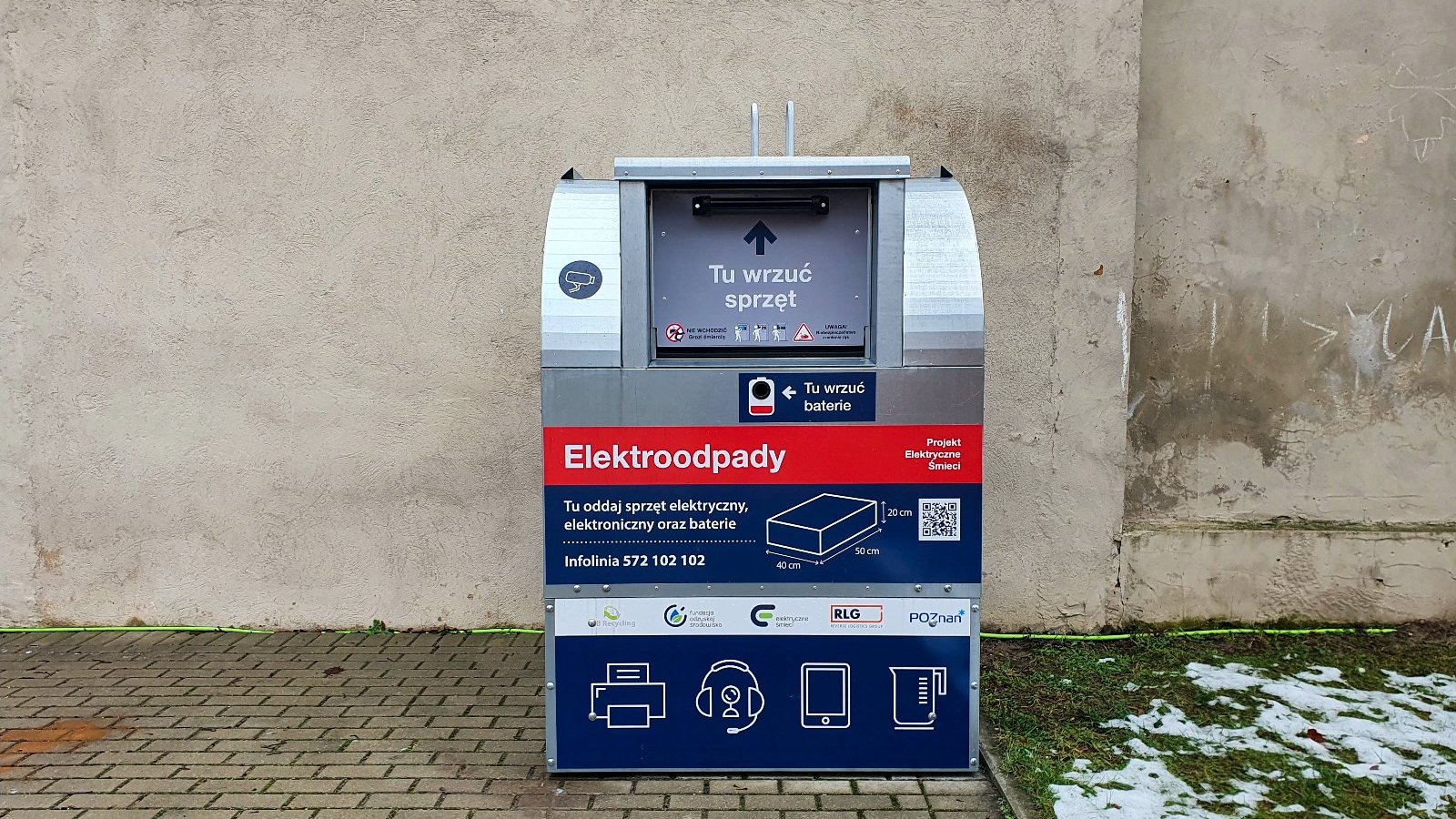 Na zdjęciu siwo-niebieski pojemnik na elektroodpady stojący na tle murku