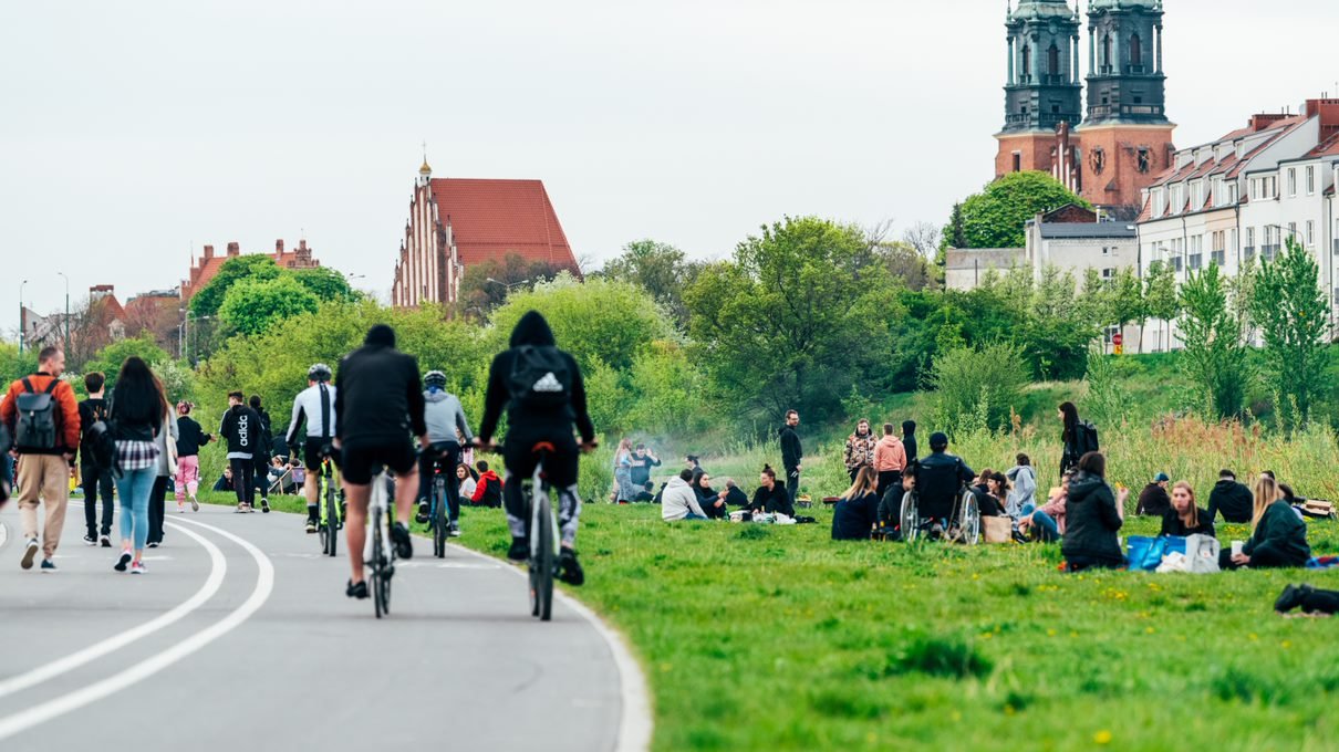 Zdjęcie przedstawia grupę ludzi wypoczywających nad Wartą. W tle wieże katedry na Ostrowie Tumskim.