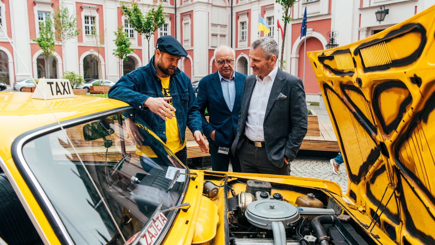 Galeria zdjęć z konferencji na dziedzińcu urzędu miasta, na zdjęciach żółta taksówka z serialu "Zmiennicy", aktor oraz zastępca prezydenta Poznania