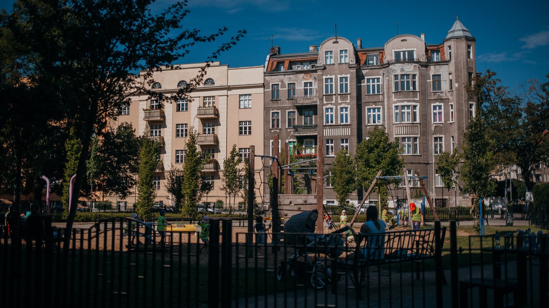 Na zdjęciu widok na kamienice, przed nimi plac zabaw oraz drzewa