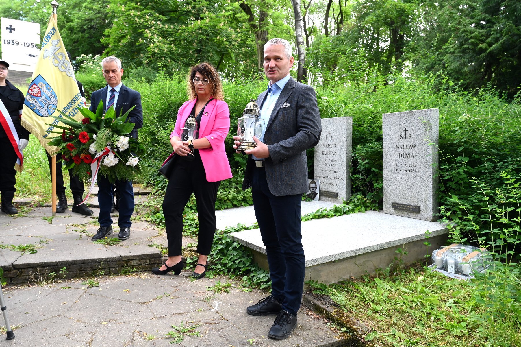 Na zdjęciu trójka ludzi stojąca przeg grobami, mężczyźni trzymają wieniec i znicz, a kobieta znicz - grafika artykułu