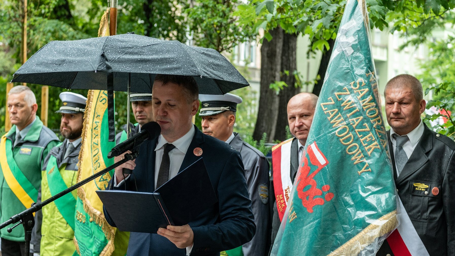 Na zdjęciu dyrektor Gabinetu Prezydenta przy mikrofonie, w tle ludzie z parasolami