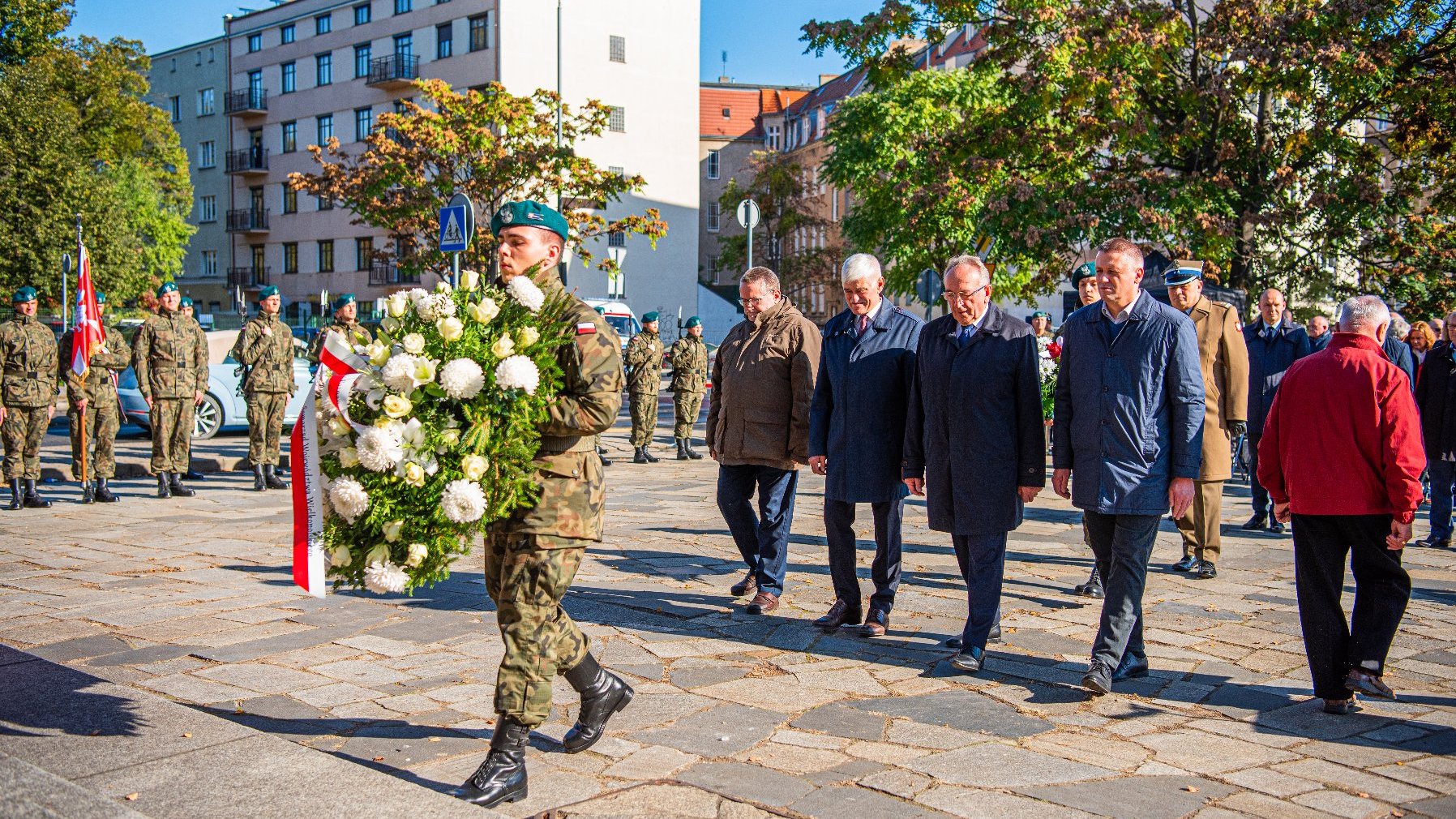 Na zdjęciu delegacja, przed nią żołnierz trzymający wiązankę