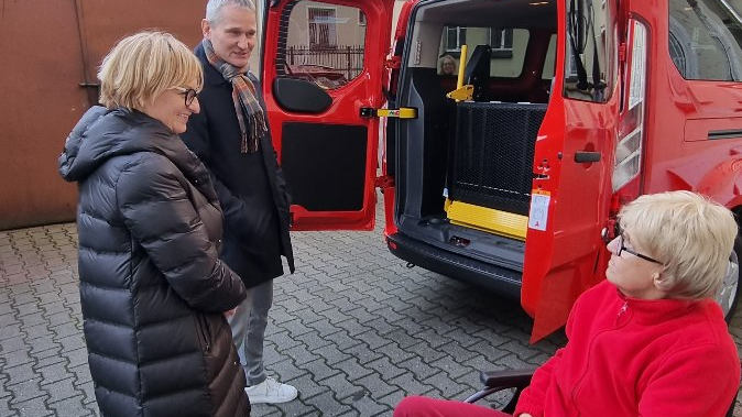 Zdjęcie przedstawia trzy osoby stojące obok czerwonego samochodu. Auto ma otwarte tylne drzwi.