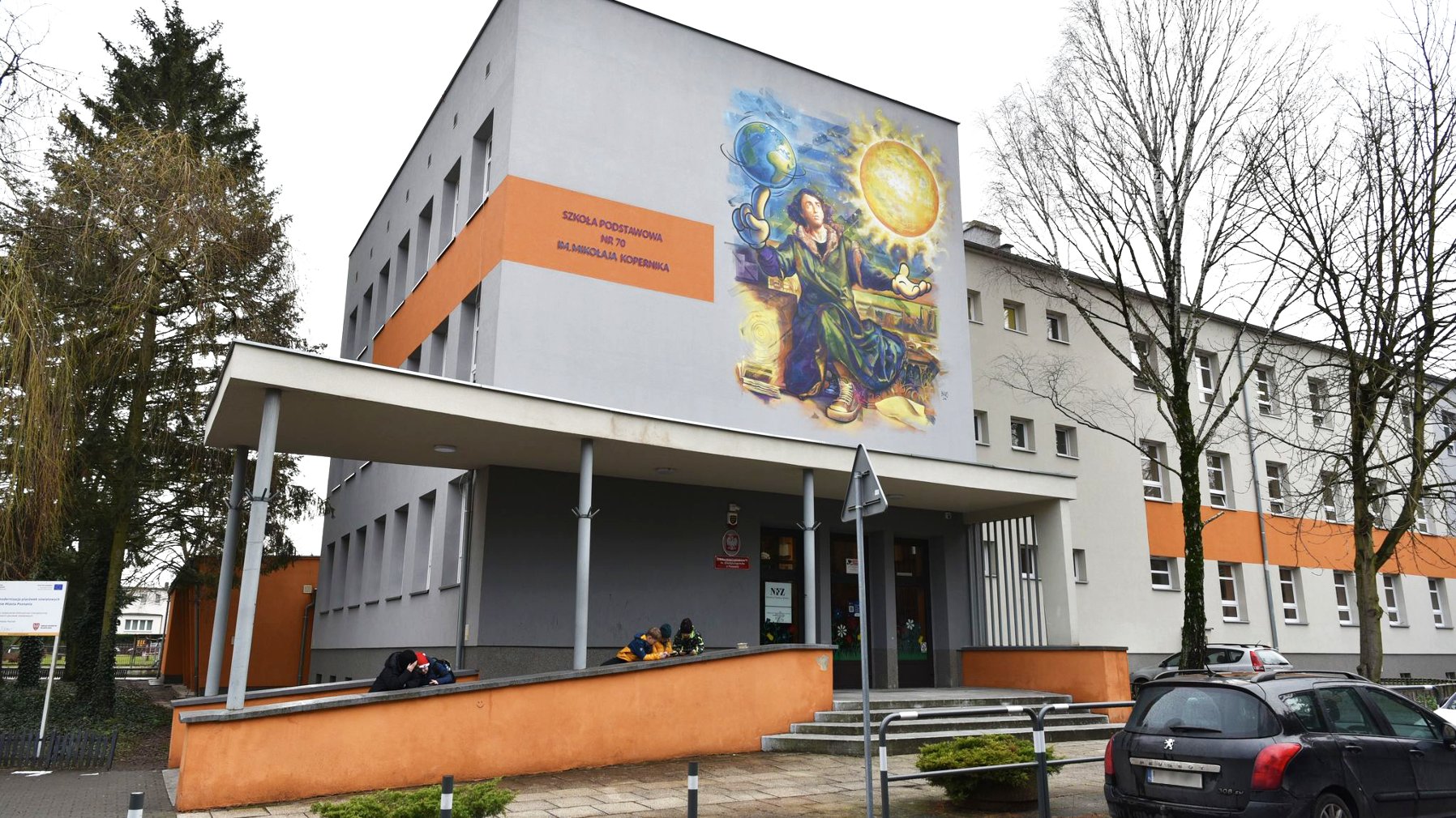 Zdjęcie przedstawia budynek szkoły. Widać na nim także uczniów oraz znajdujący się na budynku mural z podobizną MIkołaja Kopernika.