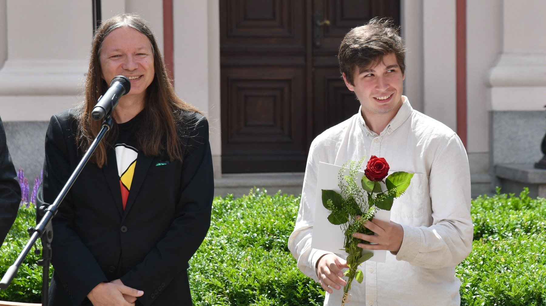 Na zdjęciu dwóch mężczyzn, jeden przy mikrofonie, drugi trzyma różę