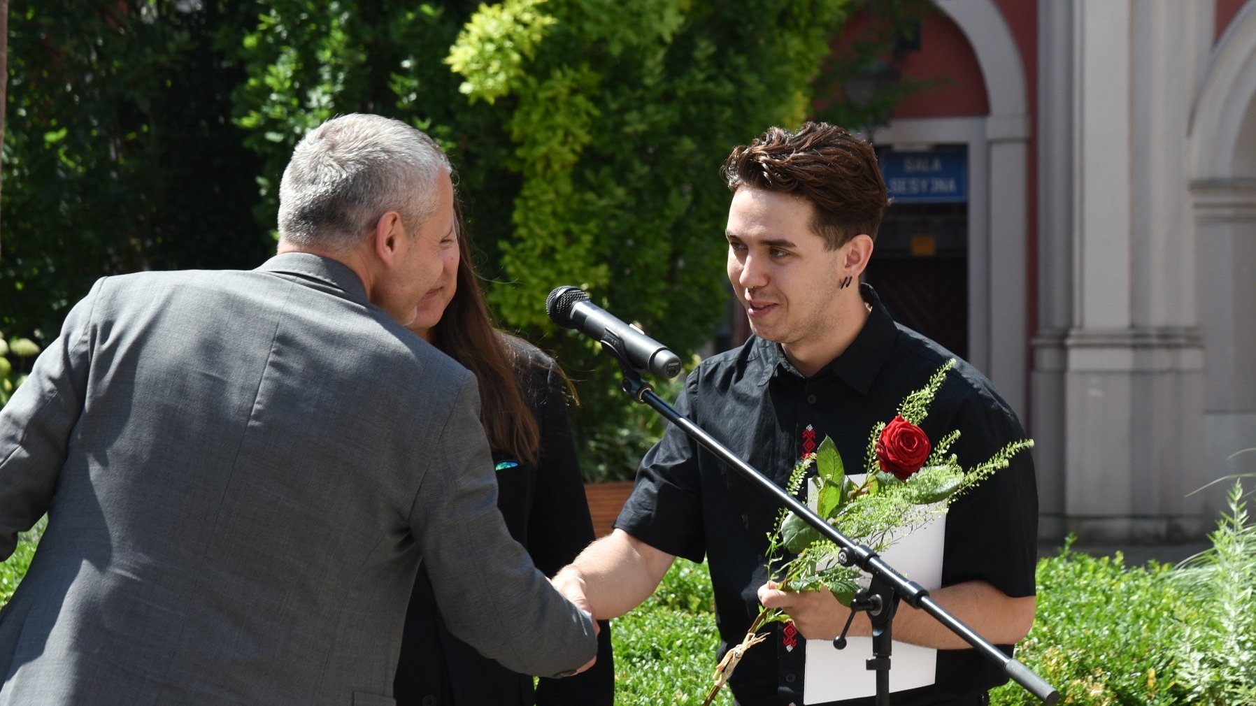 Na zdjęciu dwóch mężczyzn podających sobie ręce, jeden z nich trzyma różę