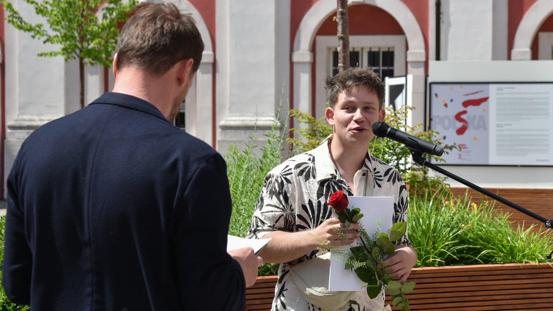 Na zdjęciu dwóch mężczyzn, jeden z nich, przy mikrofonie, trzyma różę