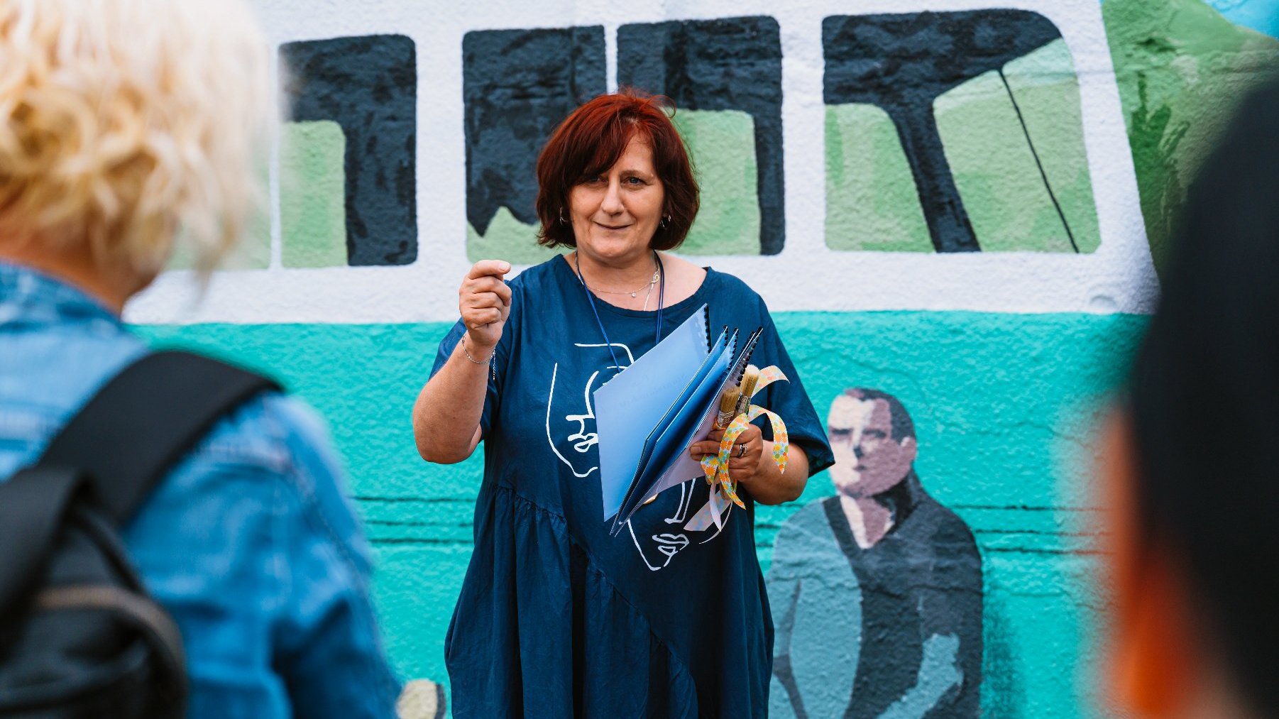 Na zdjęciu kobieta przed muralem, gestykuluje