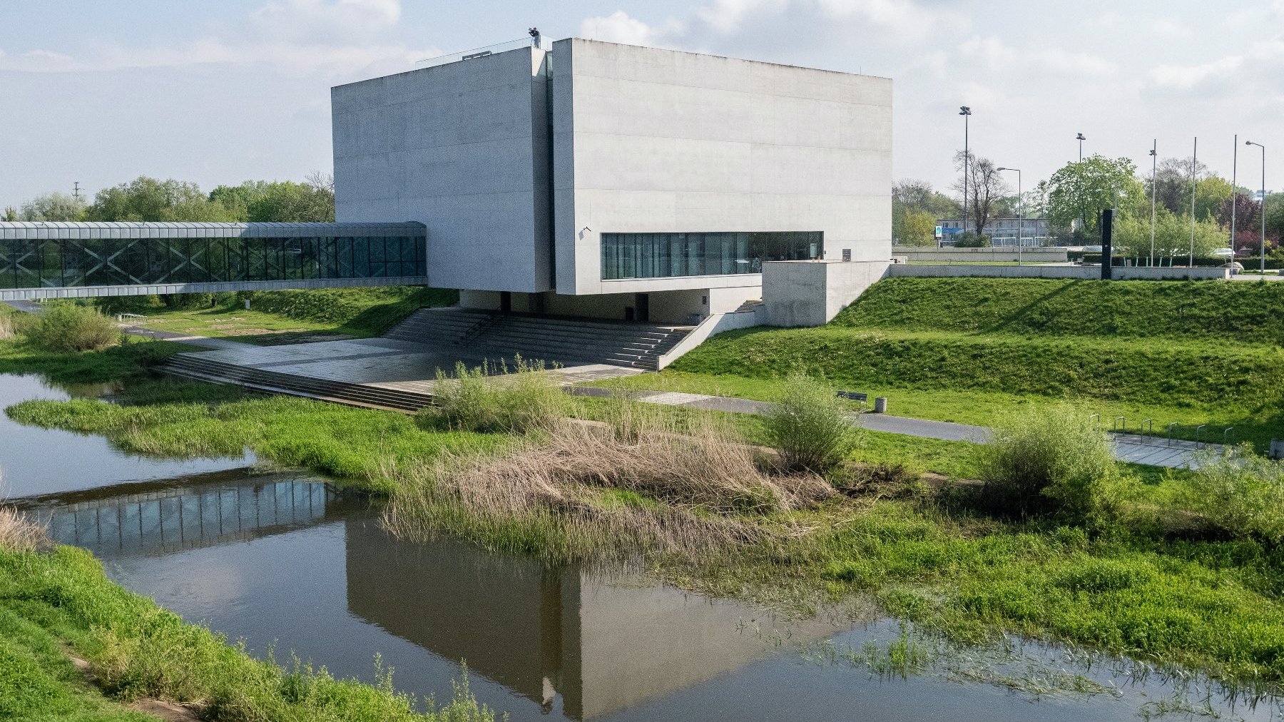Zdjęcie przedstawia geometryczny budynek "Bramy Poznania", położony nad rzeką i otoczony zielenią.