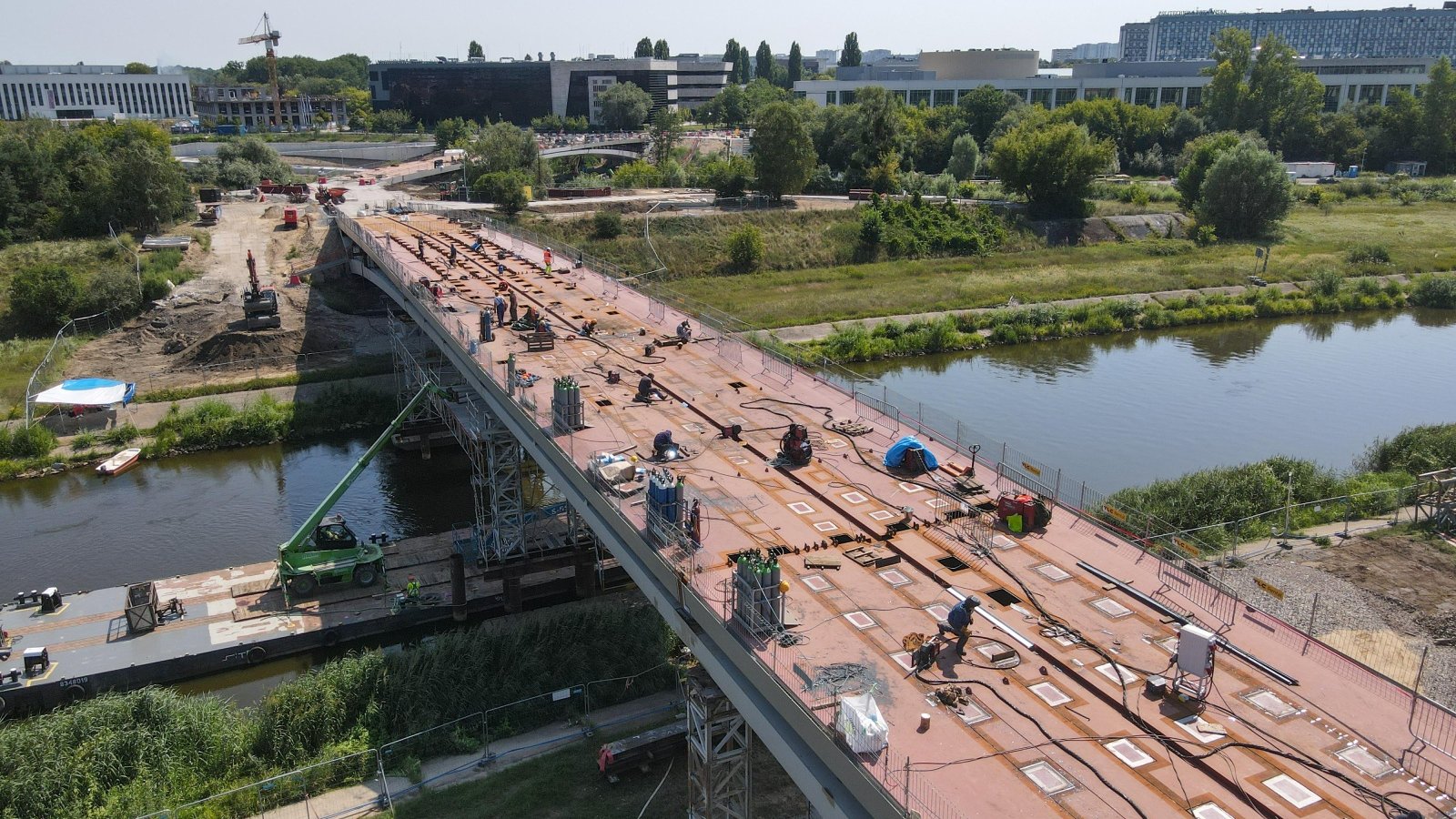 Zdjęcie przedstawia budowę mostu oraz rzekę i budynki.