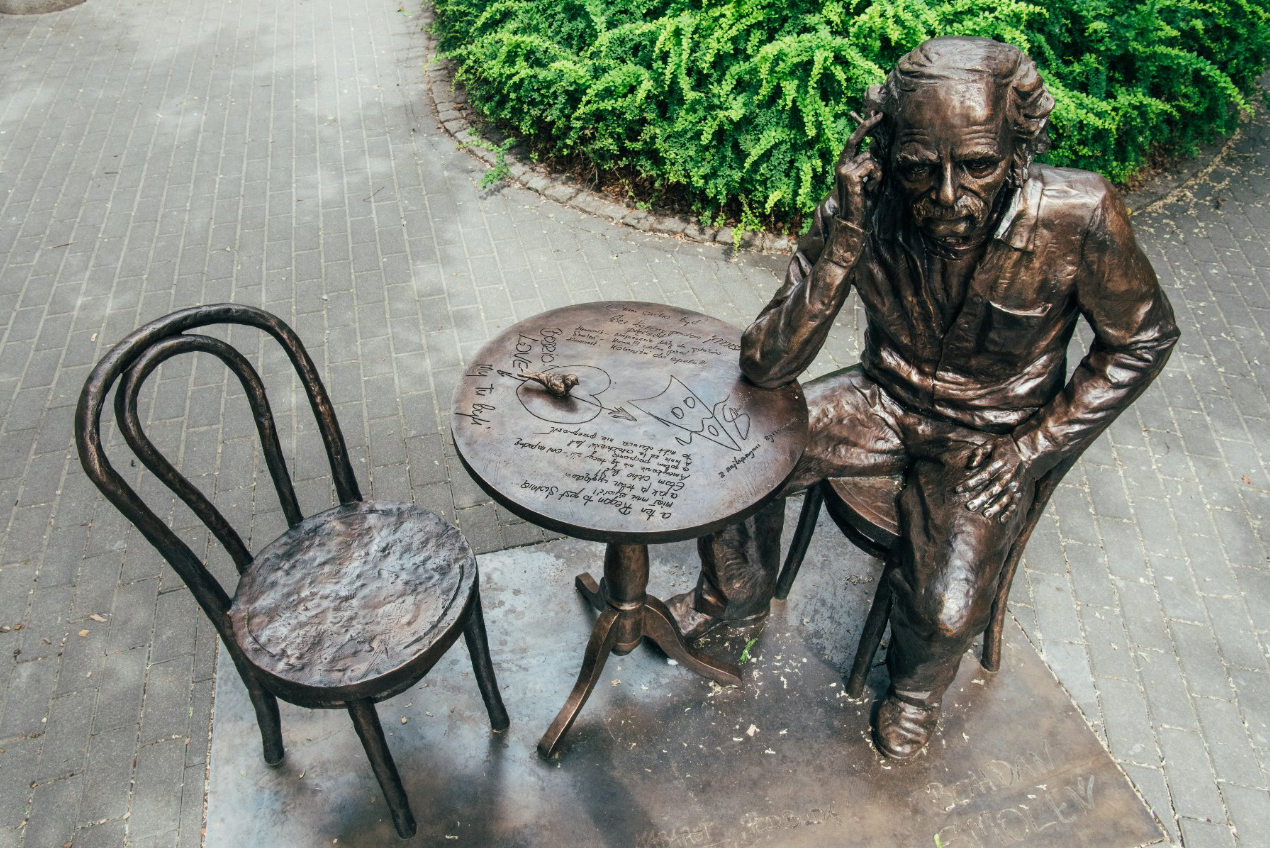 Brązowa statua osoby siedzącej przy małym okrągłym stole z krzesłem obok, znajdująca się na zewnątrz na wybrukowanym terenie z zielenią w tle. Na powierzchni stołu znajdują się różne napisy i rysunki, w tym serce z przechodzącą przez nie strzałą, kawałek pizzy oraz kilka ręcznie pisanych notatek. - grafika artykułu