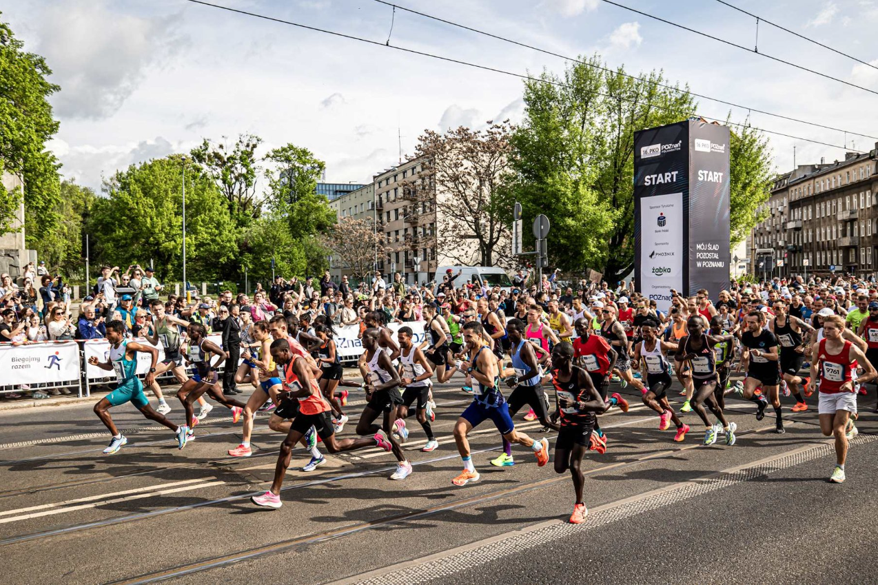 Grupa biegaczy na starcie wyścigu. Biegacze ubrani są w różnorodne stroje sportowe i uchwyceni w momencie rozpoczęcia biegu. W tle widoczni są kibice, drzewa i budynki. Na pierwszym planie duży czarno-biały baner z napisem 'START', wskazujący początek wyścigu. Scena jest dynamiczna i pełna energii, z różnorodną grupą uczestników i widzów. - grafika artykułu