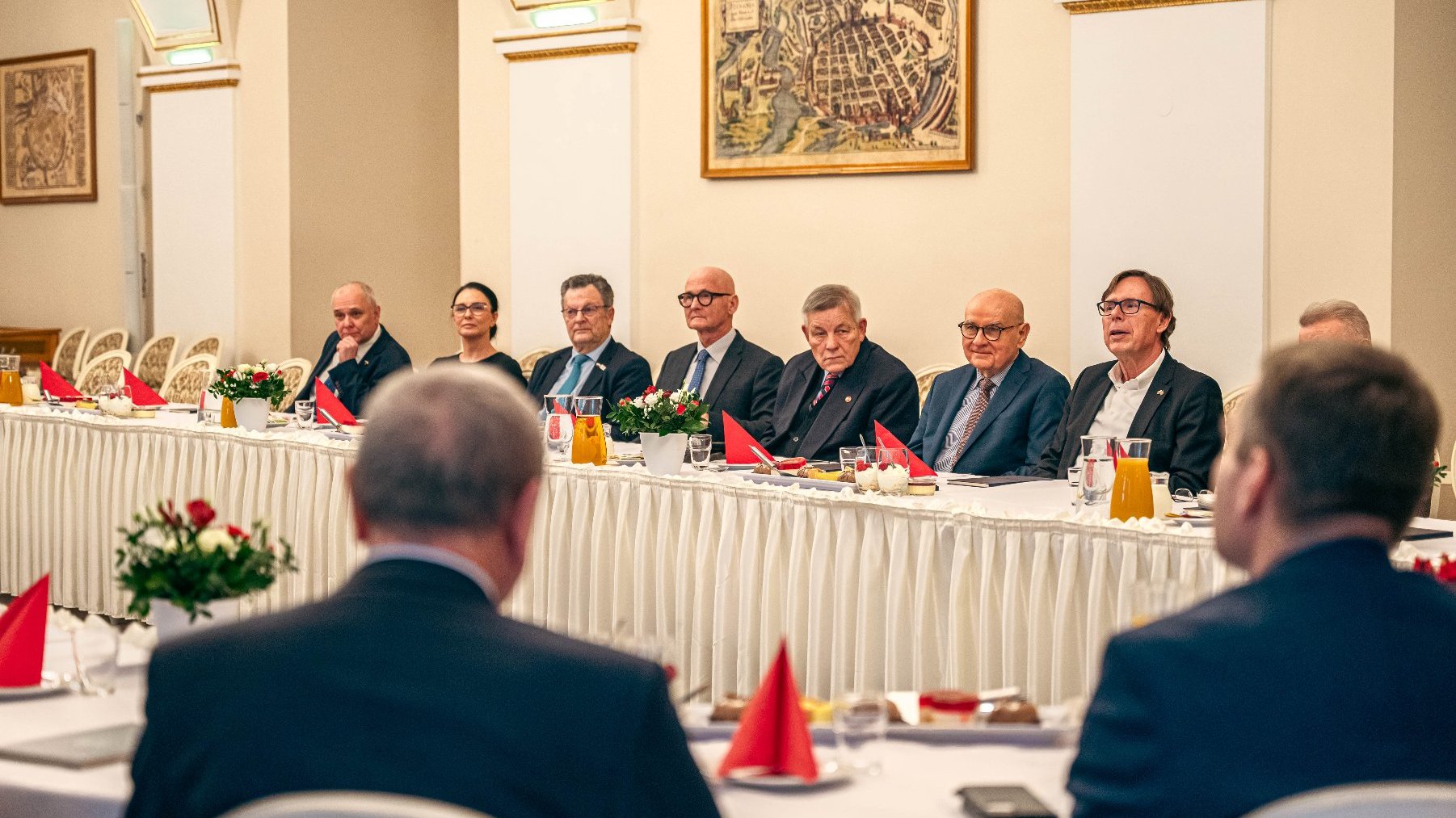 Zespół ludzi w formalnym spotkaniu w eleganckim pomieszczeniu. Stoł z białym fartuchem, czerwonymi ręcznikami, kwiatami i dzbankami z napojami. Osoby w formalnych strojach, rozmawiające lub słuchające prezentacji.