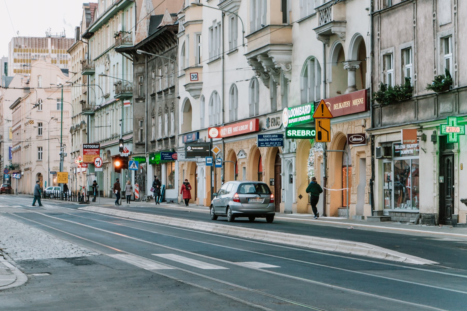 Obraz przedstawia widok ulicy w obszarze miejskim z kilkoma budynkami i sklepami wzdłuż ulicy. Ludzie spacerują po chodniku, a kilka samochodów jest widocznych na drodze. Budynki mają różne tablice i reklamy, w tym zielony krzyż na farmacji, tablica z napisem "LOMBARD" na antykwarii i inne tablice reklamowe. Ulica ma torowiska tramwajowe i przycisk do przejścia dla pieszych. - grafika artykułu