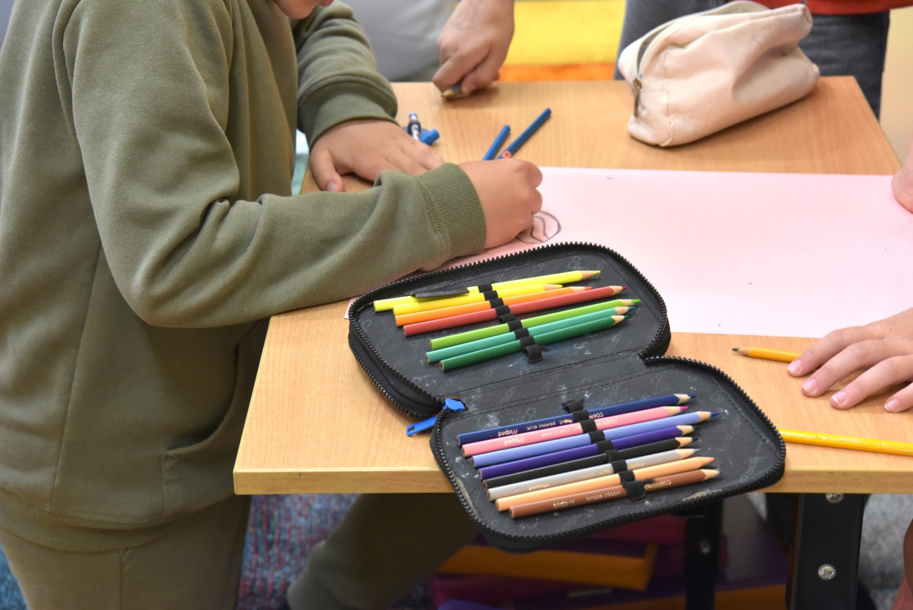Muzeum Literackie Henryka Sienkiewicza zaprasza dzieci i ich opiekunów na warsztaty artystyczne Dziecko w zielonej bluzie rysujące na różowej kartce papieru. Na drewnianym stole leży otwarty piórnik z zestawem kolorowych kredek. W tle widać fragmenty rąk innych osób.