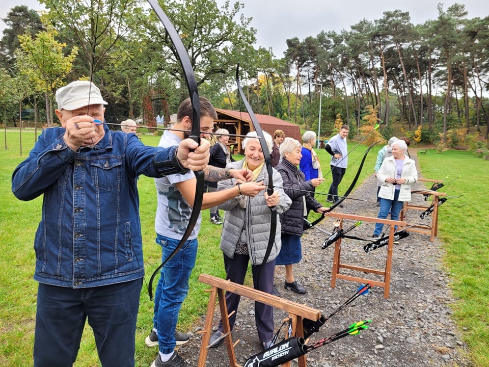 Teren zielony, w oddali drzewa. Mężczyzna strzela z kuszy. Obok młodszy mężczyzna pokazuje kobiecie, jak strzelać z kuszy. Kilka osób przygląda się temu. - grafika artykułu