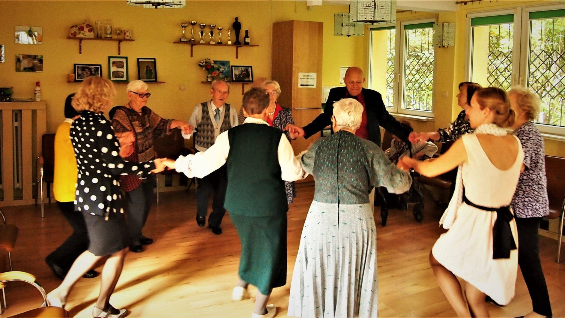 Pomieszczenie. Mężczyźni i kobiety trzymają się za ręce, tworzą kółko i tańczą.