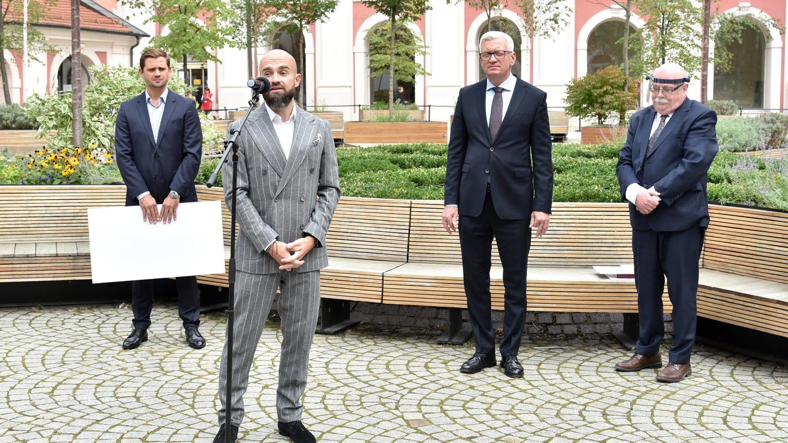 Dziedziniec urzędu miasta. Czterej mężczyźni w garniturach, na pierwszym planie, przy mikrofonie Sebastian Kulczyk. W tle drzewa