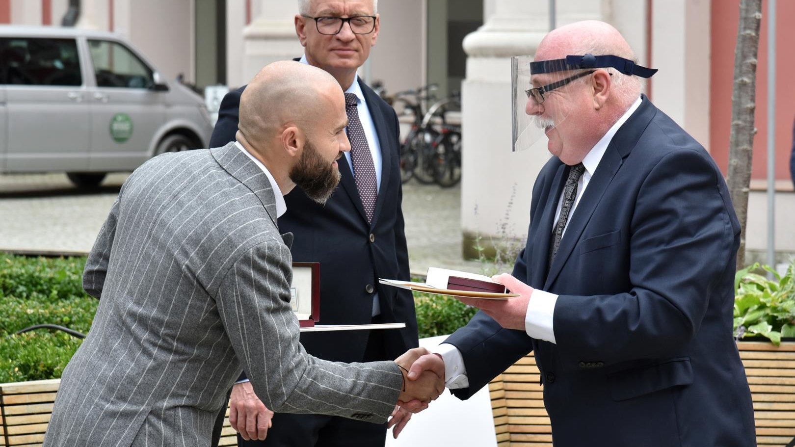 Dziedziniec urzędu miasta. Sebastian Kulczyk i dr Daszkiewicz podają sobie dłonie. Profesor w dłoni trzyma medal. Za nimi stoi Jacek Jaśkowiak, prezydent miasta