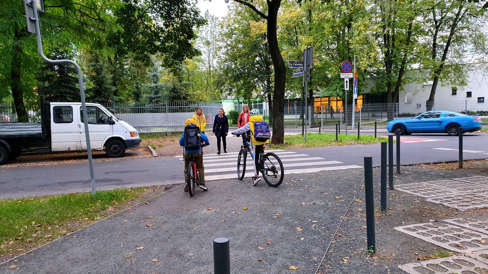 Dzieci przejeżdżają ulicę na rowerach. 3 mamy wracają do aut po odprowadzeniu dzieci