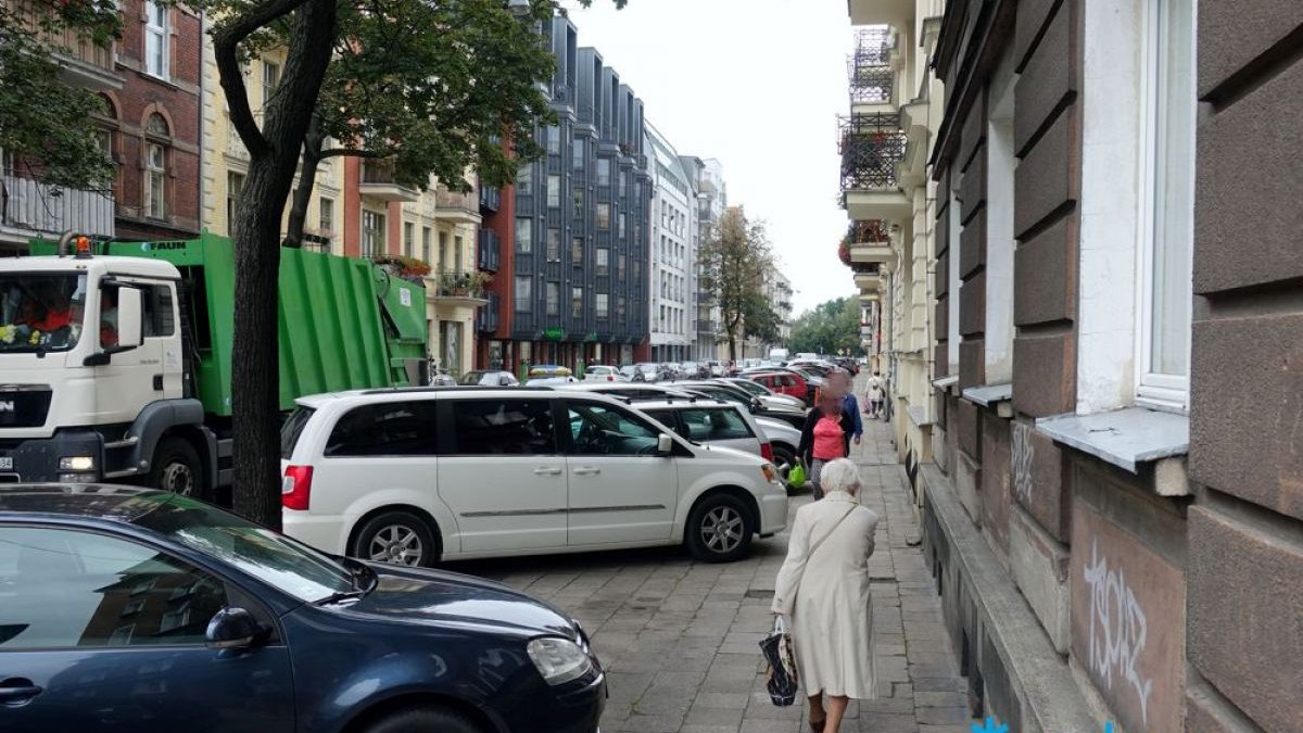 Zatłoczona ulica. Samochody parkują na chodniku, po którym idzie starsza pani. Na ulicy - śmieciarka