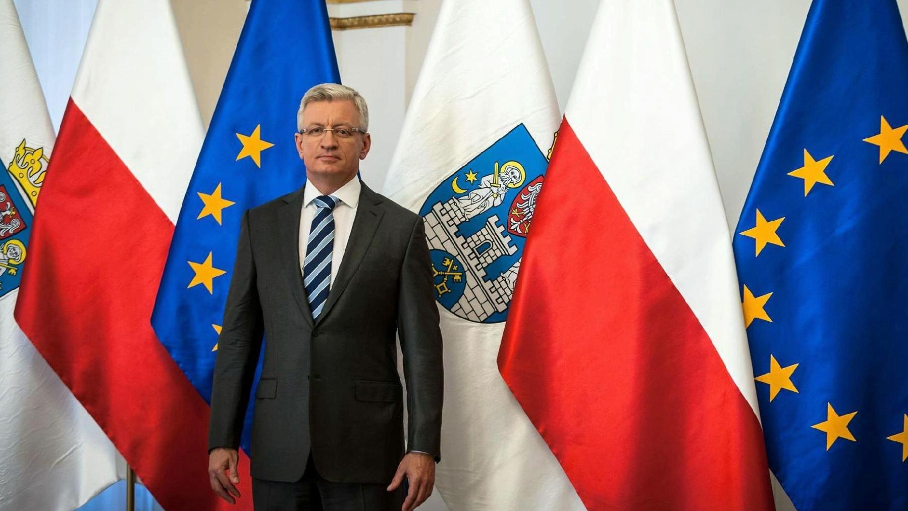 Zdjęcie przedstawia prezydenta Poznania, Jacka Jaśkowiaka na tle flag (miasta Poznania, Polski i Unii Europejskiej). - grafika artykułu