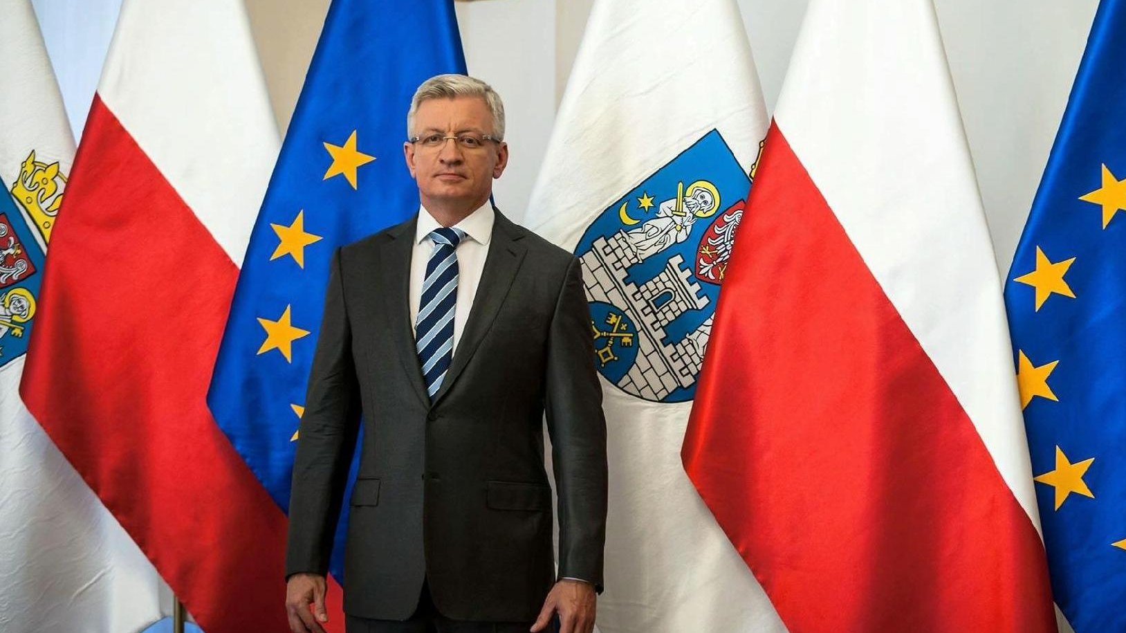 Zdjęcie przedstawia prezydenta Poznania, Jacka Jaśkowiaka na tle flag (miasta Poznania, Polski i Unii Europejskiej).