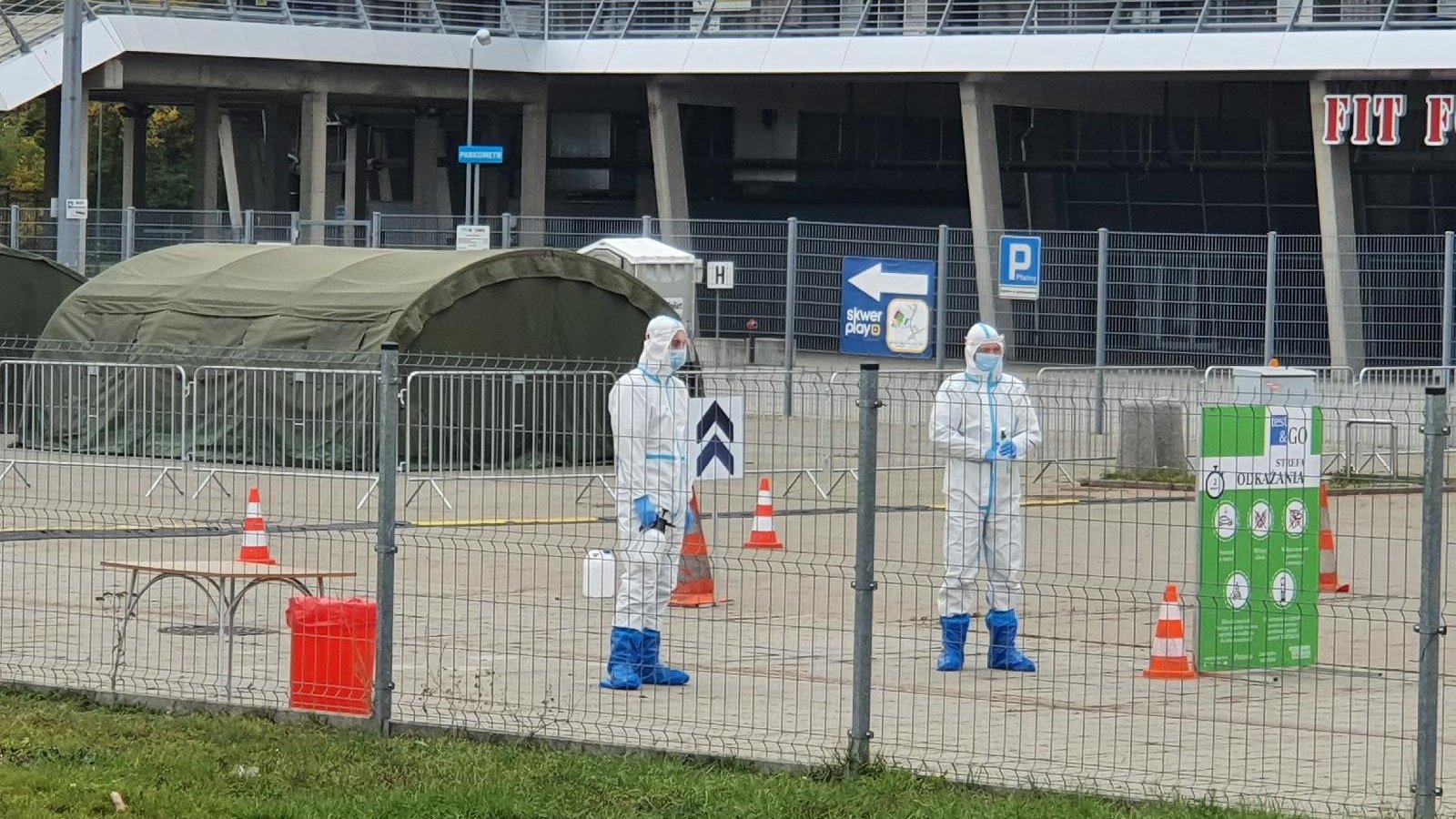 Dwie osoby w kombinezonach stoją przy miejskim stadionie.