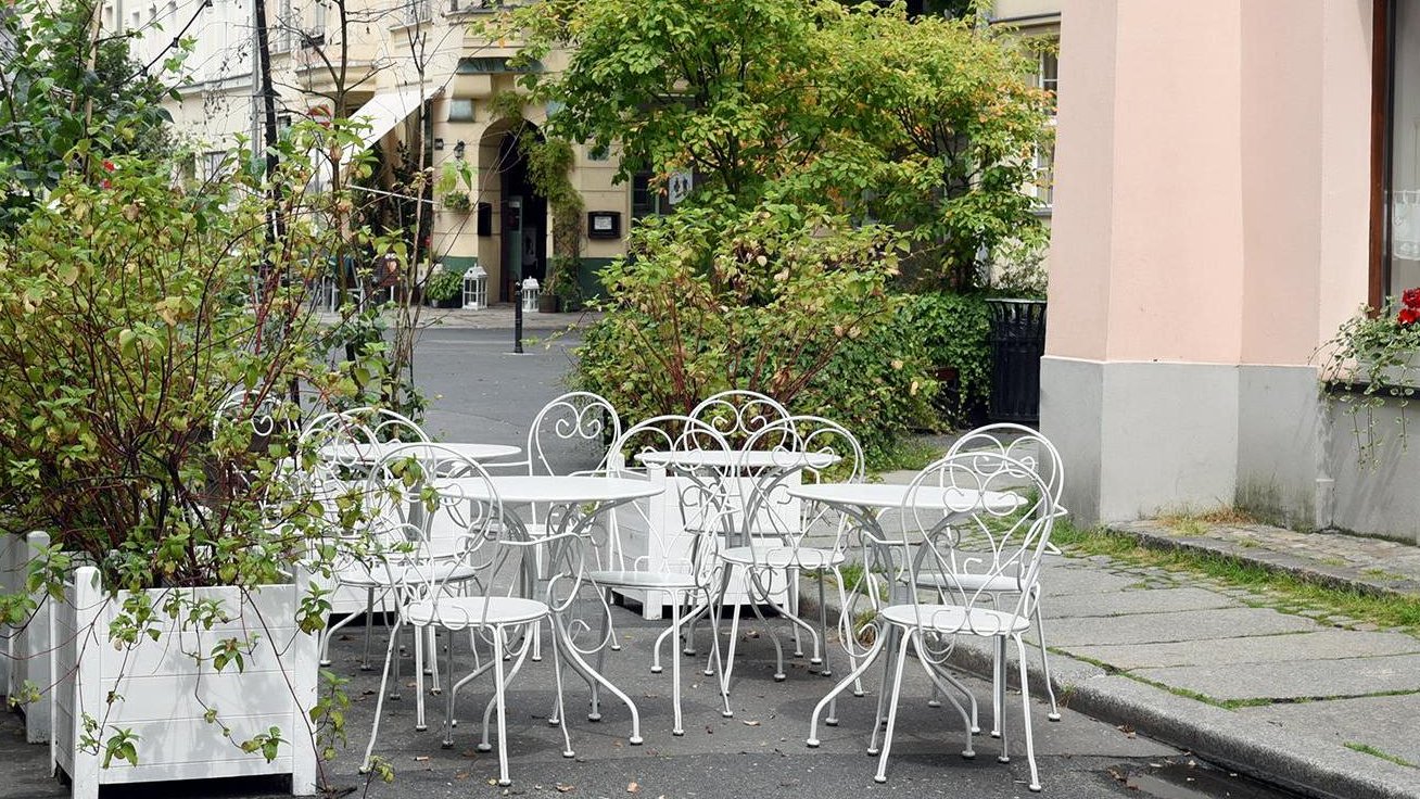 Zdjęcie: ogródek gastronomiczny na Starym Mieście, obok kamienicy stoją białe stoły i krzesła, wokół nich rośliny w białych donicach